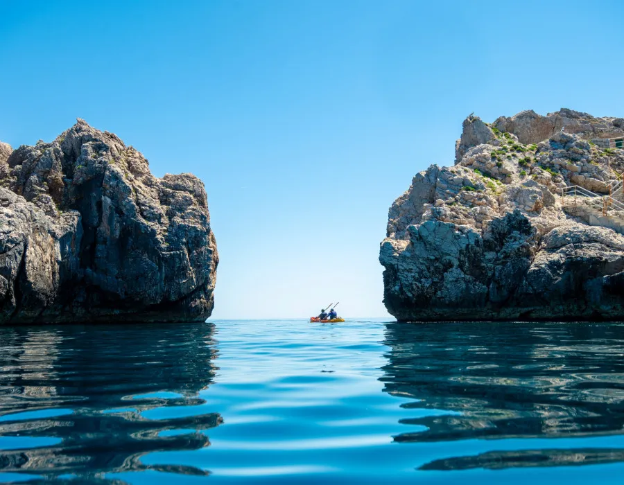 Two participants kayak through an opening in the rocks on clear, calm water and underneath a bright blue sky