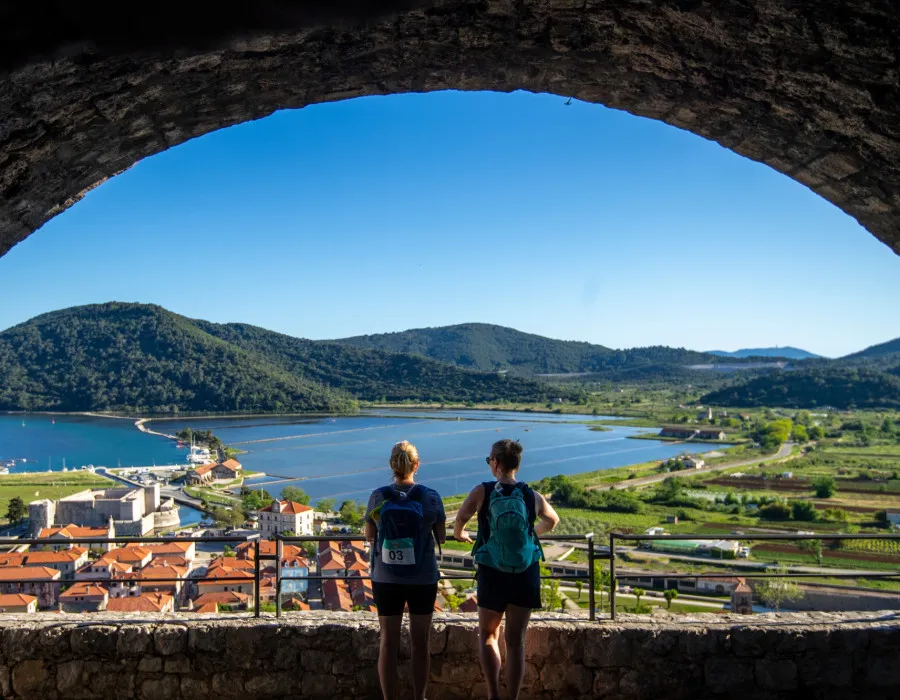 Two women stand under an arch, looking out onto buildings, water and greenery