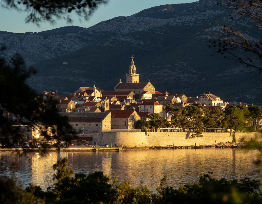 Buildings enclosed by a stone wall sit beneath mountainous scenery, with water stretching around