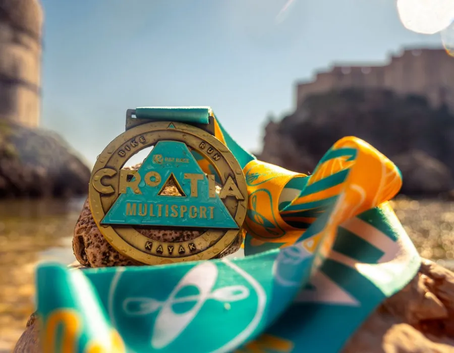 A Croatia Multisport medal with a colourful ribbon rests on rocks beside the water, with sunlit buildings blurred in the background