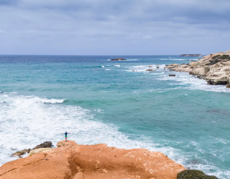 A lone participant stands on an orange rock with arms outstretched, looking out across the pale blue sea