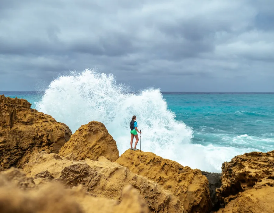 Woman standing on a sandy rock, with the sea splashing against it