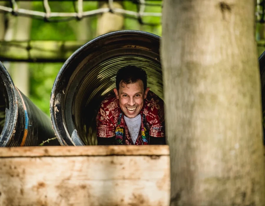 A smiling man, wearing a colourful shirt, crawling through a tunnel in a forest