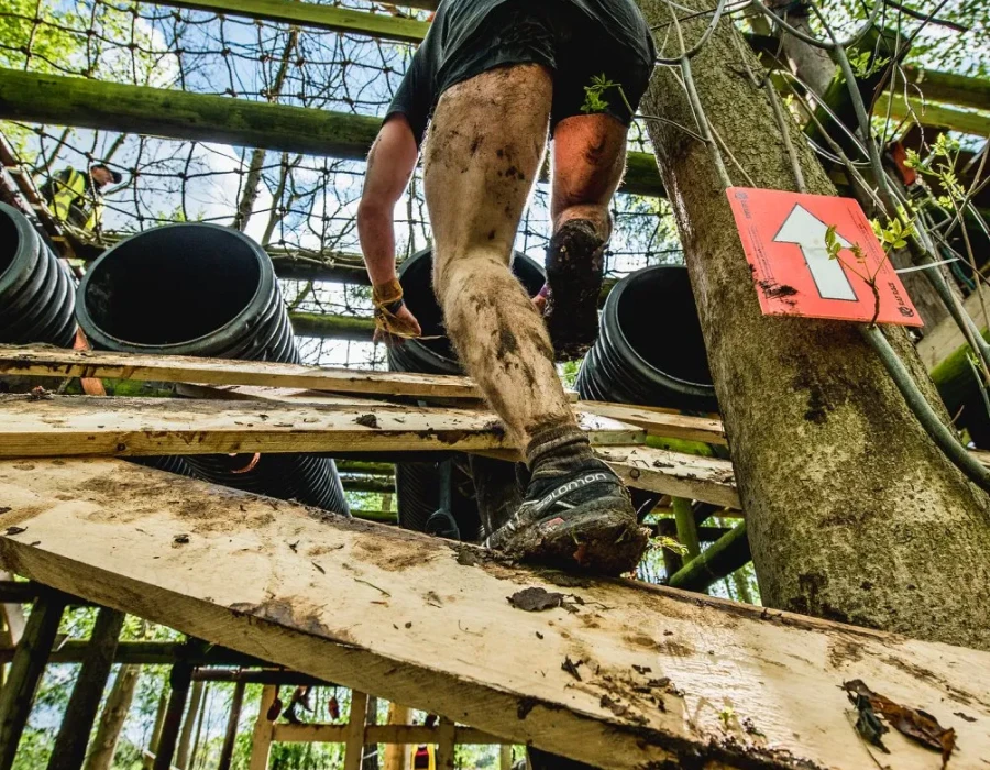 A mud-covered man, climbing up planks of wood on an obstacle course
