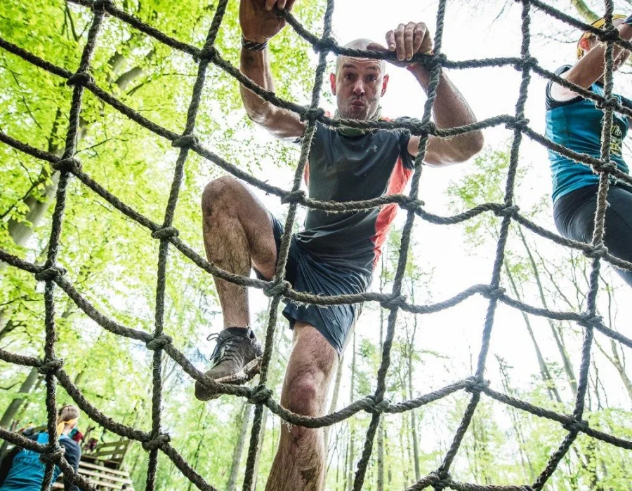 A mud-covered man climbs up a rope net obstacle