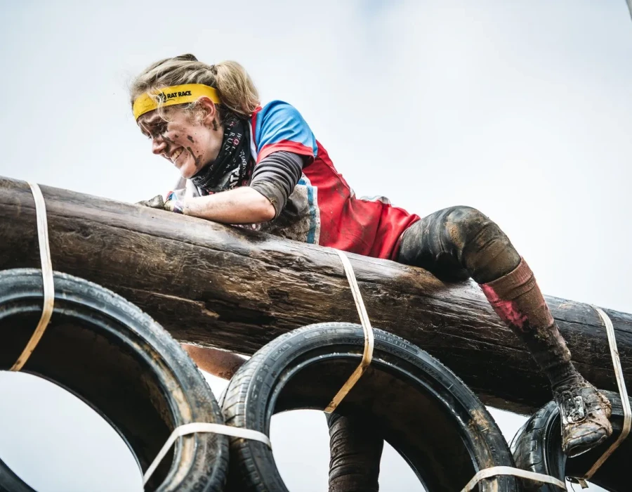 Woman, covered in mud, holding onto a wooden pole above tyres