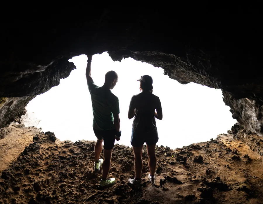 Two participants stand inside a rocky cave entrance, silhouetted against the bright daylight outside
