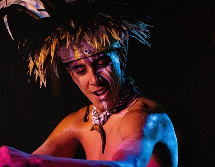 A performer wears a feathered headdress, face paint, and shell necklaces under dramatic coloured lighting