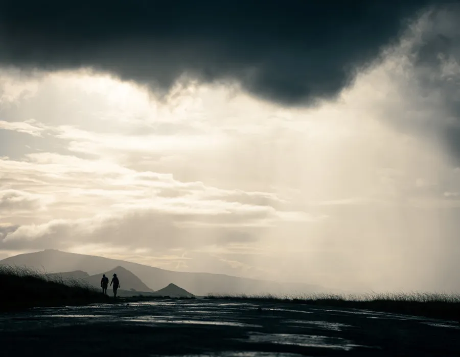 Two silhouettes walk across a grassy field with mountains in the background and the dark sky above