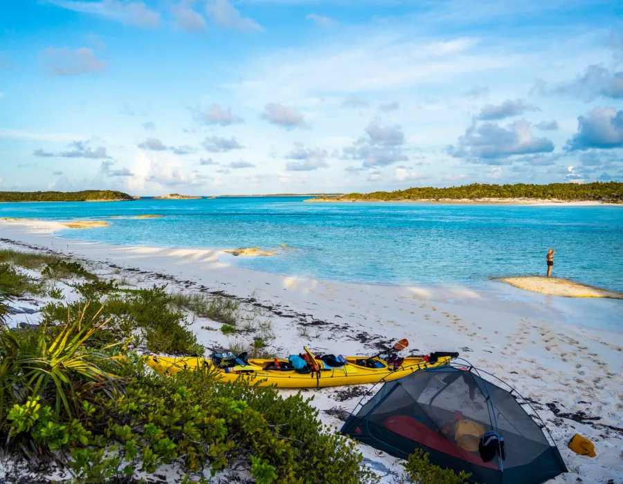 Man stood by the water on a sandy beach with a kayak and tent behind him