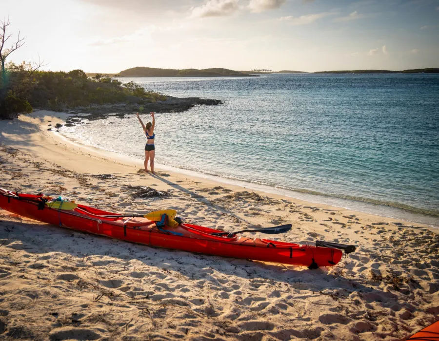 Woman standing infront of a colourful kayak on a sandy beach with her arms in the air