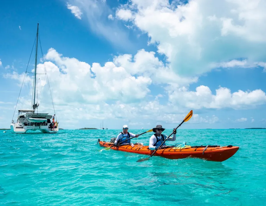 Participants kayaking through clear water with a boat behind them and sunny sky above