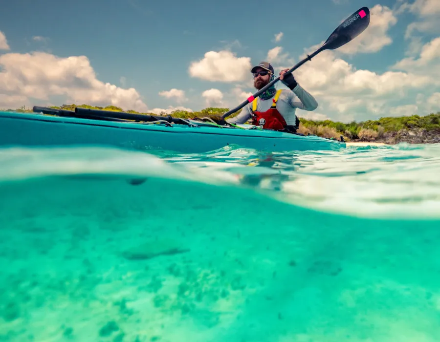 Man kayaking through clear, shallow water with greenery behind him
