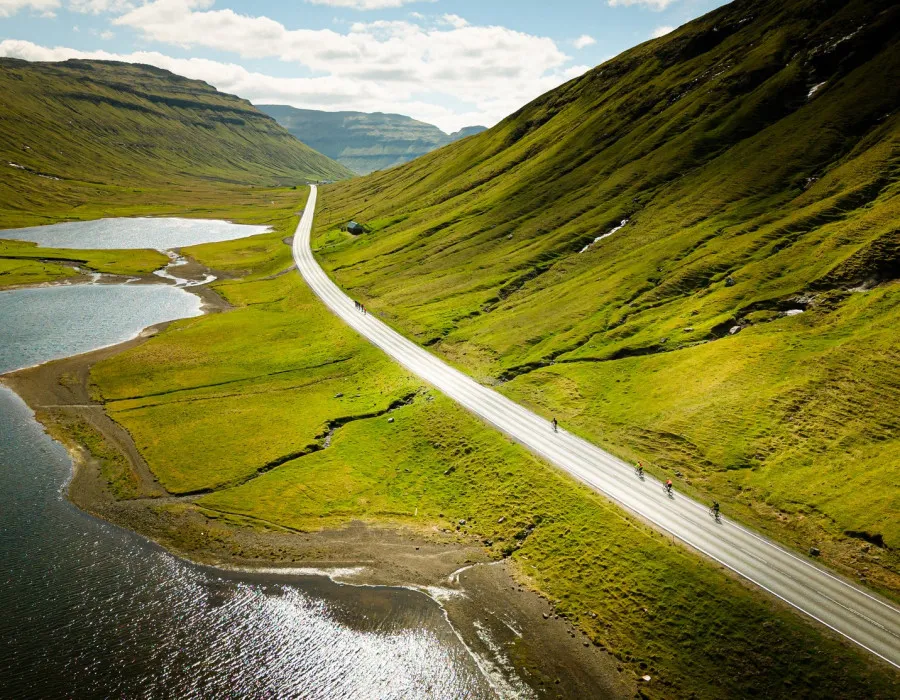 Cyclists riding along a straight road, surrounded by water and green mountains