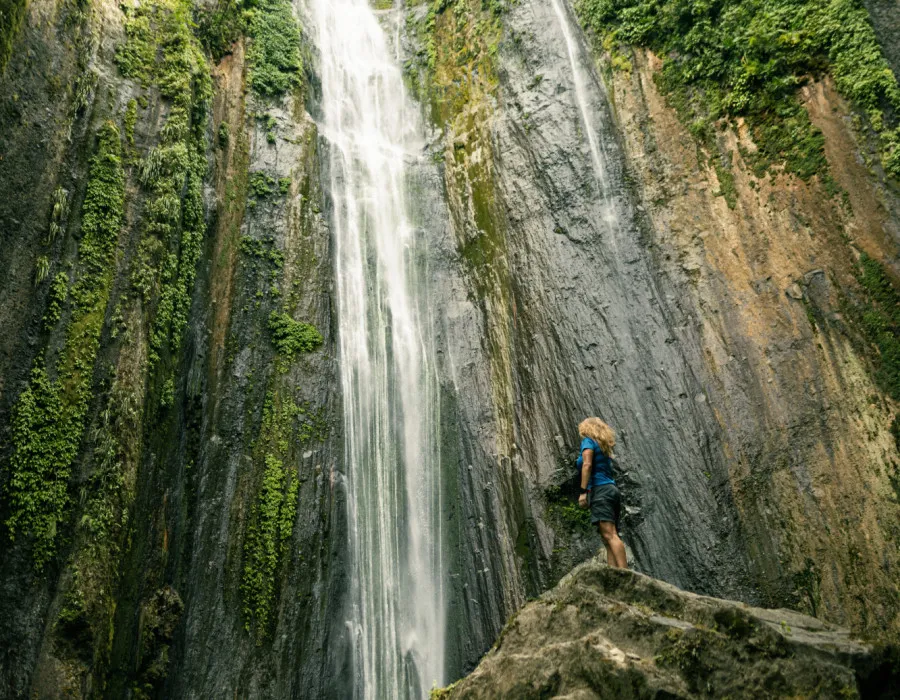Woman stood on jagged rock, looking up to waterfalls, surrounded by greenery