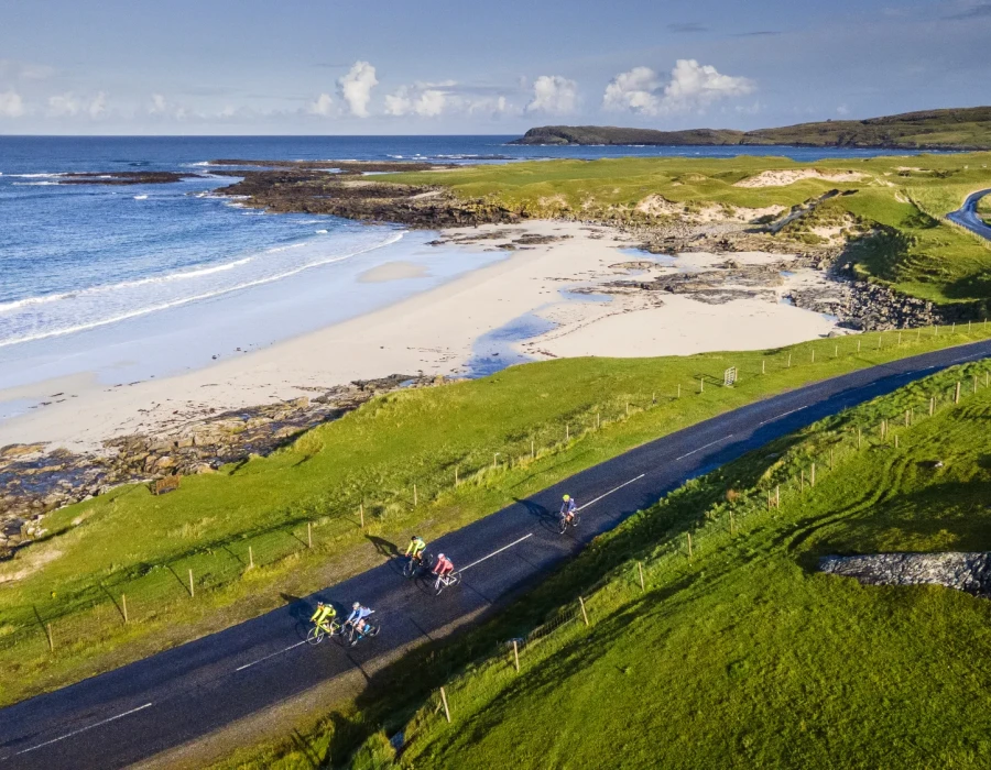 Cyclists riding along coastal road, with sandy beach to one side and green field to the other