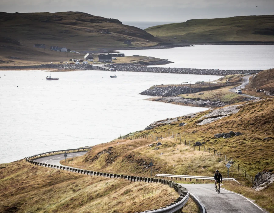 Single cyclist riding along road with fields either side and water behind