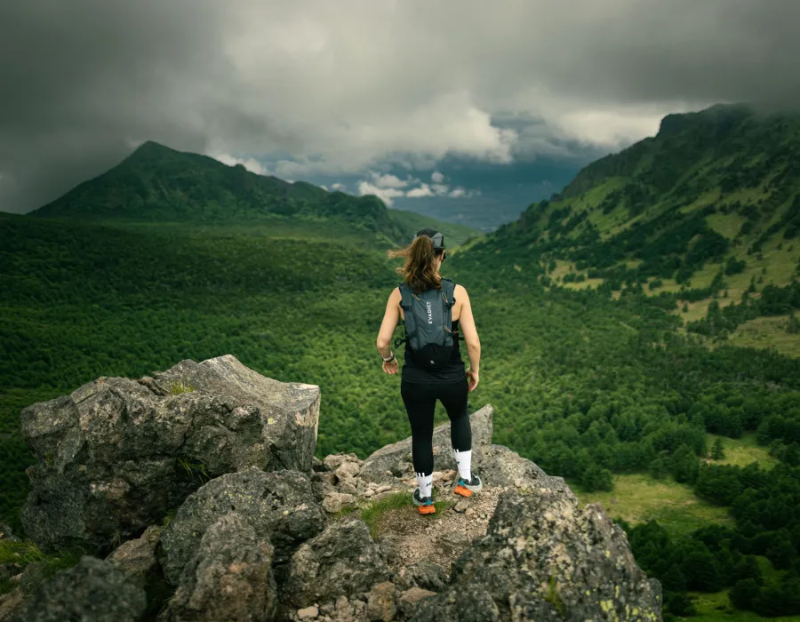 Woman stood on rock, looking out to forest and mountains with a cloudy sky above