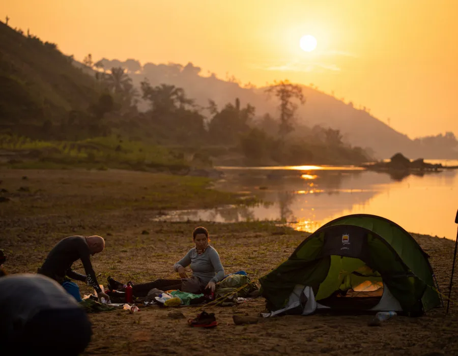 Participants sat near their tent infront of water at sunset with trees and mountains in the background