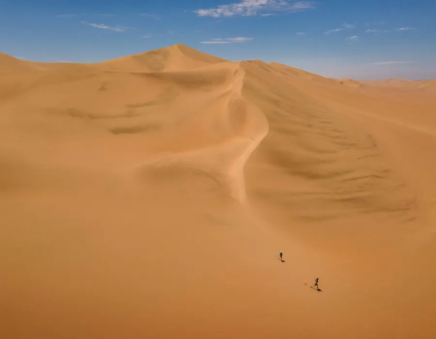 Two people running down smooth sand dunes with bright sky above