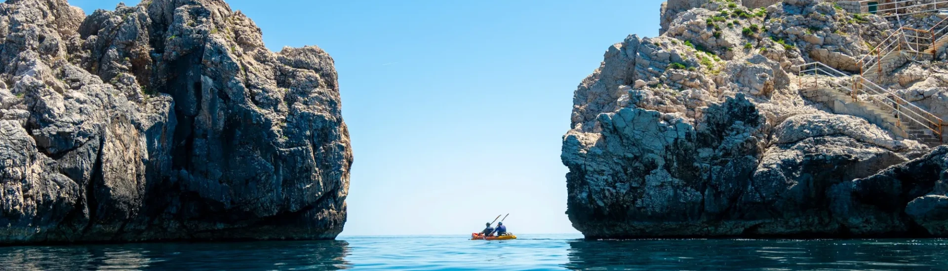Two participants kayak through an opening in the rocks on clear, calm water and underneath a bright blue sky