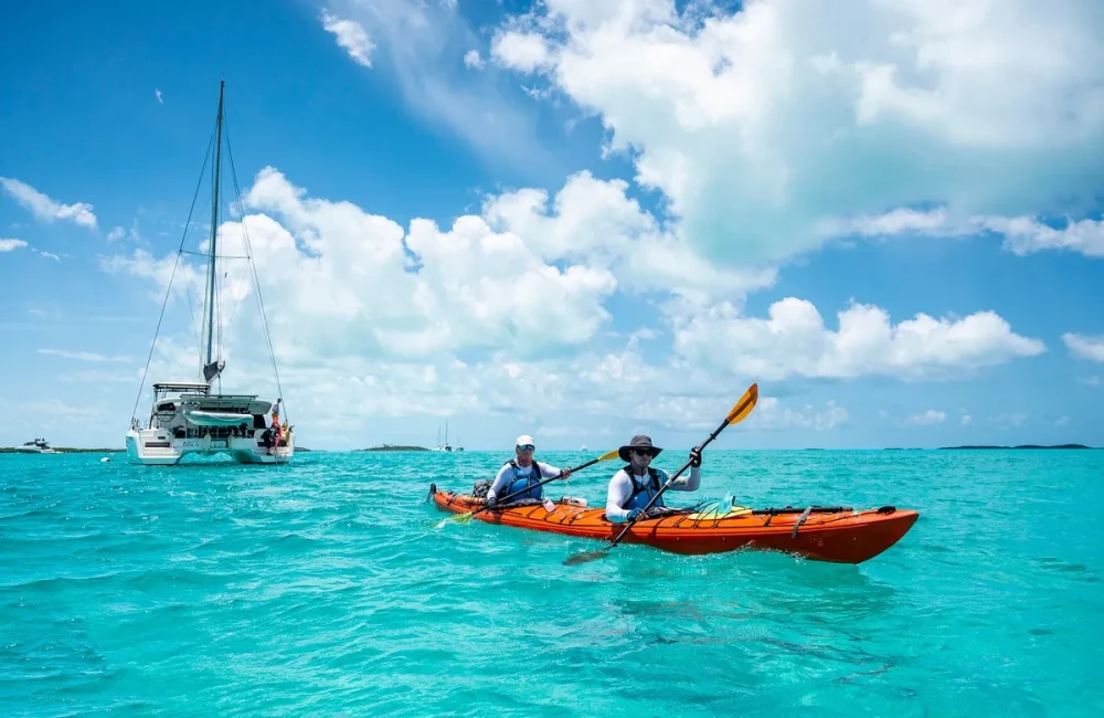 Participants kayaking through clear water with a boat behind them and sunny sky above