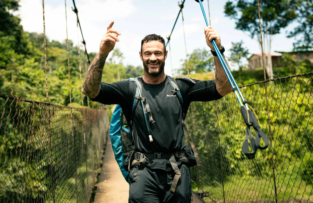 Smiling man with his hands and trekking poles in the air walking over bridge