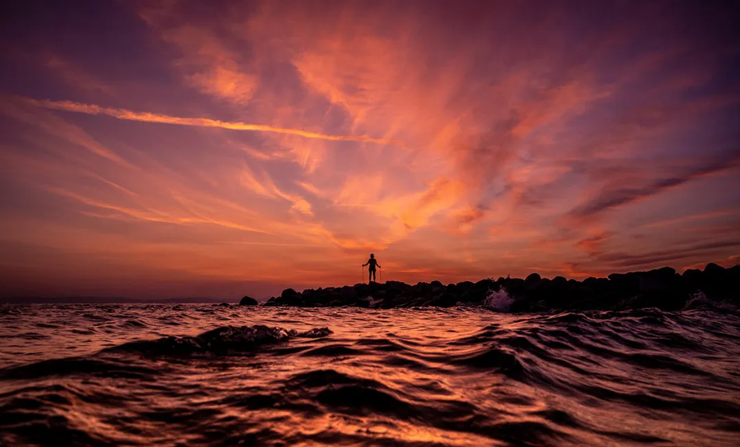 Person stood on rocks by sea and colourful sky above