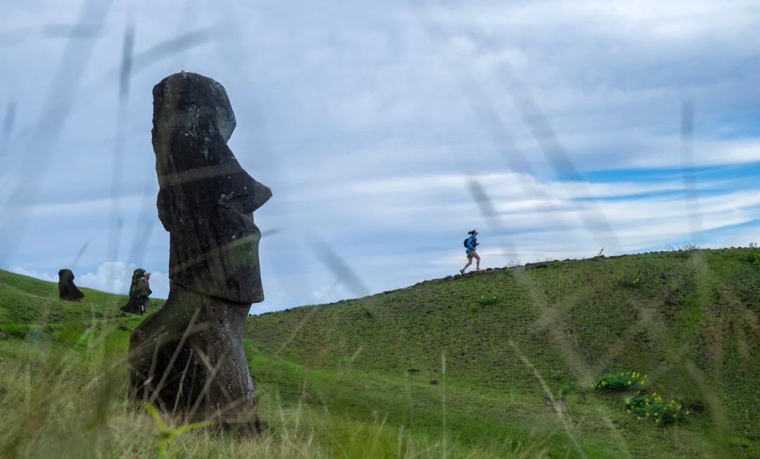 A large statue and woman running over a grassy hill with blue sky above her
