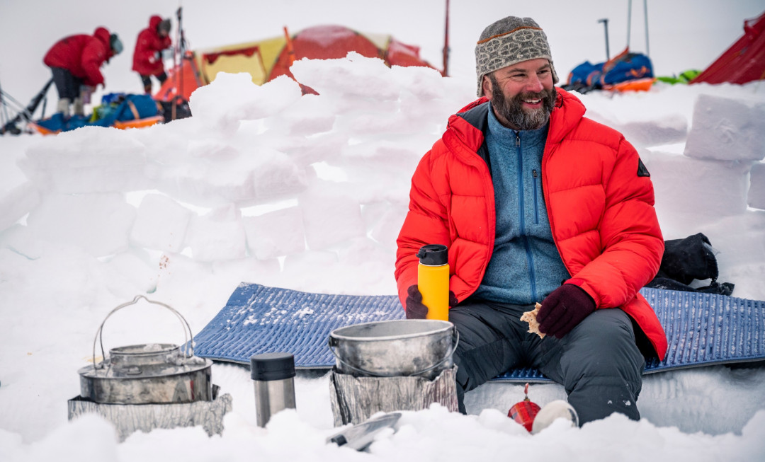 Man sat on snow, wearing a bright coloured coat and smiling
