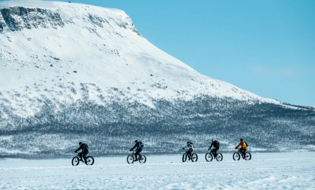 Group of cyclists riding fat bikes across a snow-covered landscape beneath a towering mountain and clear blue winter sky.