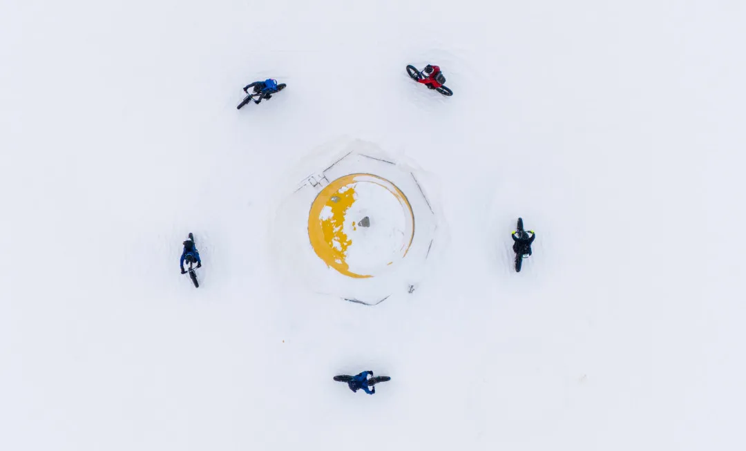 Aerial view of five cyclists riding fat bikes in a circle around a snow-covered yellow structure in a vast white winter landscape