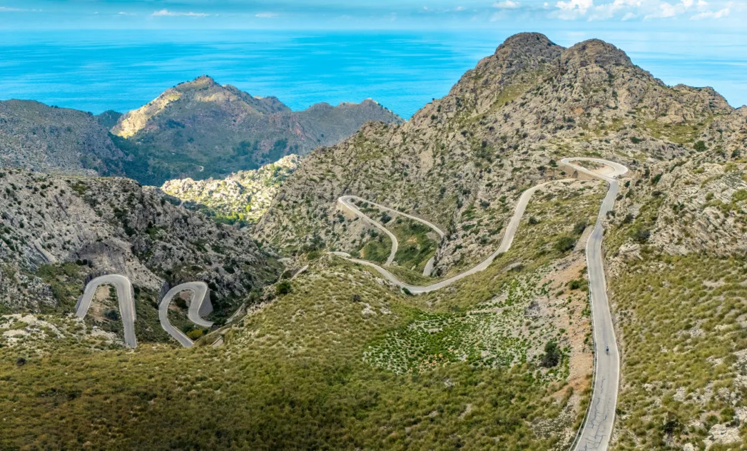 A winding road snakes through rugged, rocky terrain dotted with patches of greenery beneath a bright blue sky