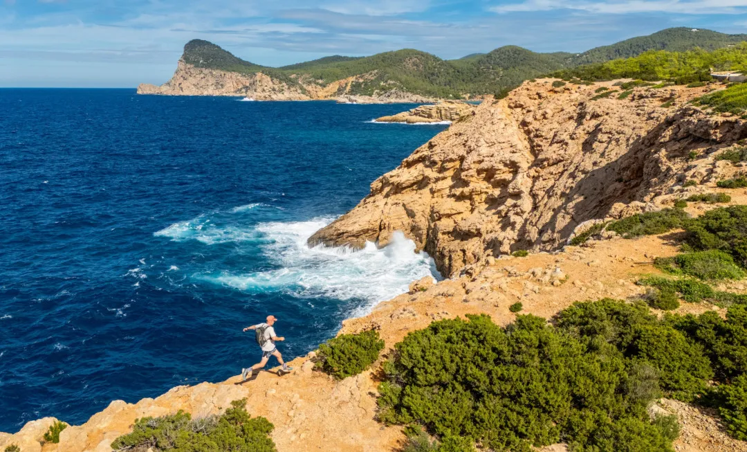 An event participant runs along the edge of a cliff beside a deep blue sea, with orange-red rock, patches of greenery, and a sunny sky above