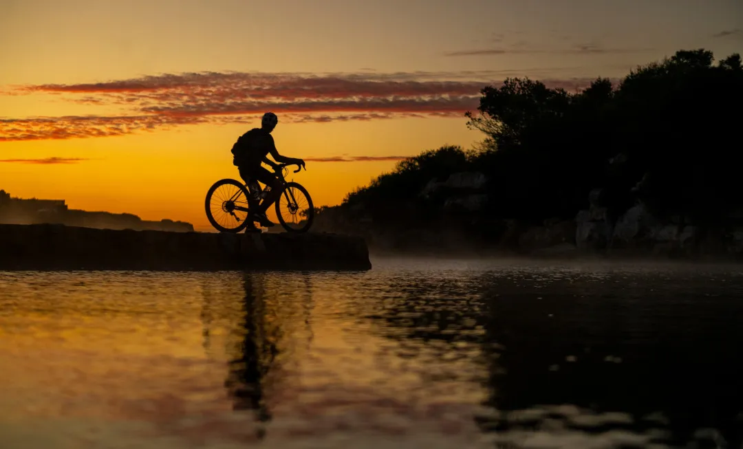 Silhouette of a lone cyclist, who has stopped by the water to admire the orange sky behind them