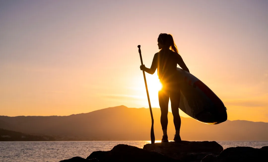 A woman stands with a paddle board, looking onto water and a pink and orange sunlit sky