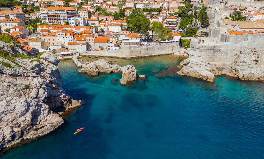 Kayaks glide across blue water, moving away from historic city walls and orange-roofed buildings