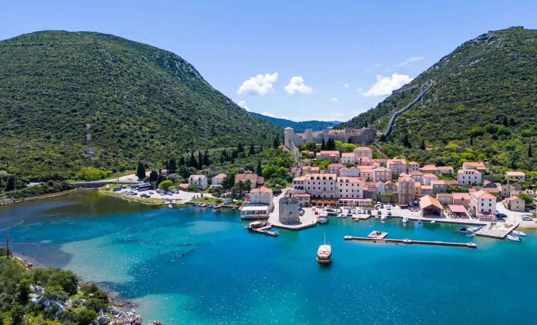 Turquoise blue water stretches towards lush green hills, with boats and waterfront buildings lining the shore in the background