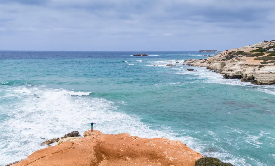 A lone participant stands on an orange rock with arms outstretched, looking out across the pale blue sea