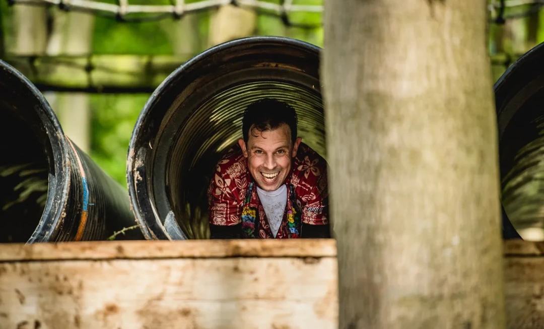 A smiling man, wearing a colourful shirt, crawling through a tunnel in a forest