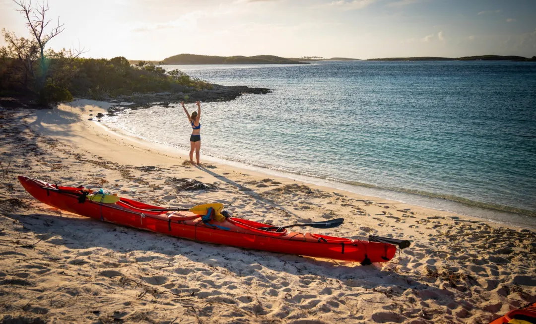 Woman standing infront of a colourful kayak on a sandy beach with her arms in the air