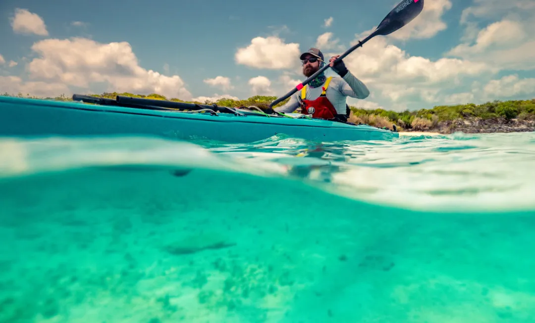 Man kayaking through clear, shallow water with greenery behind him