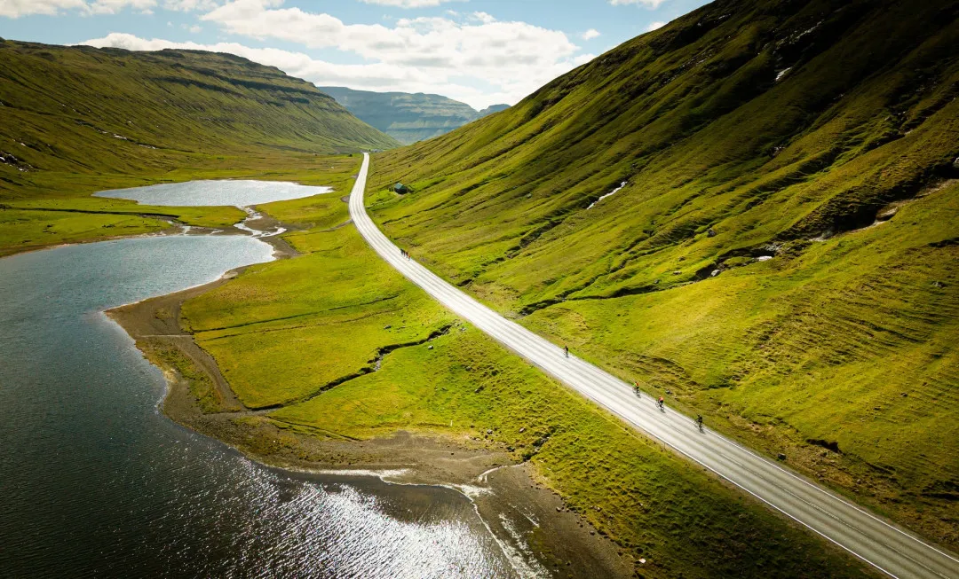Cyclists riding along a straight road, surrounded by water and green mountains