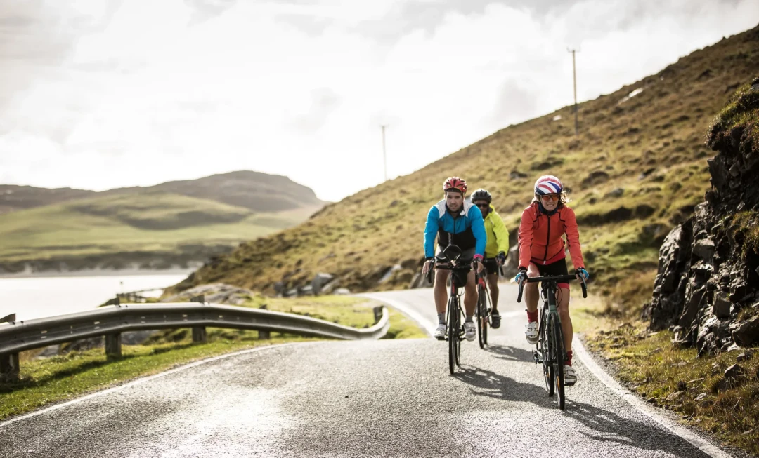 Three cyclists riding along a road with rocky, grassy terrain behind