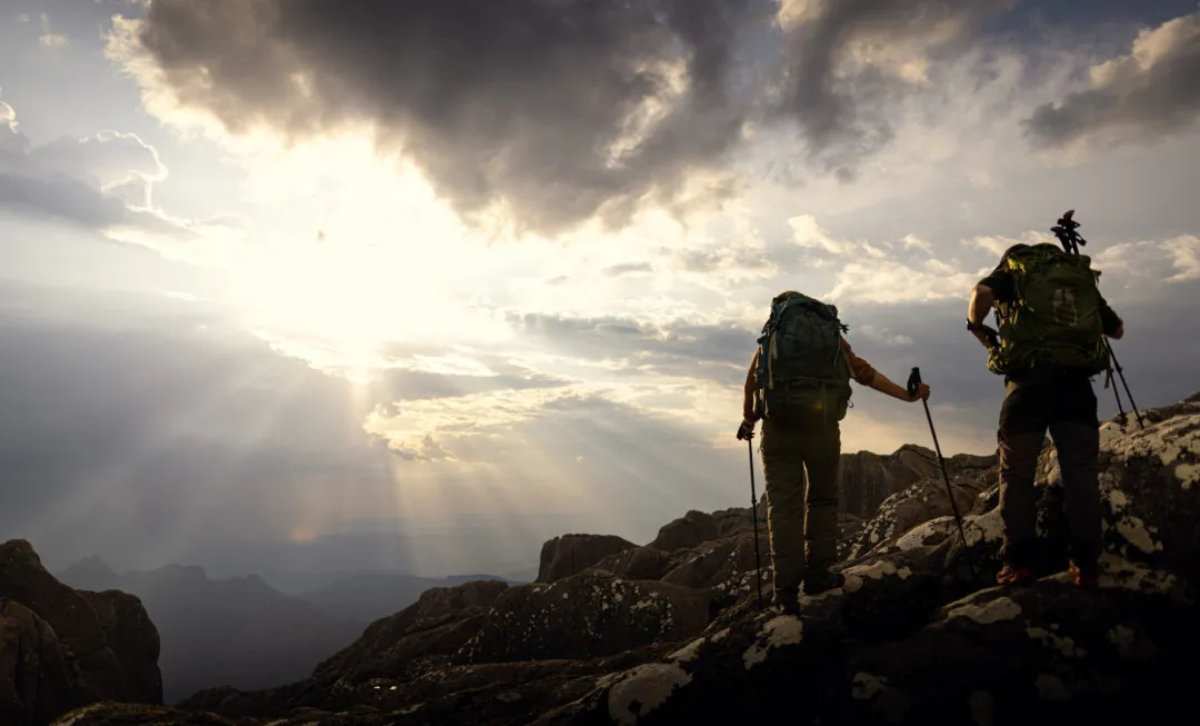 Two people, wearing rucksacks and using trekking poles climbing over rocky terrain with bright sky above them