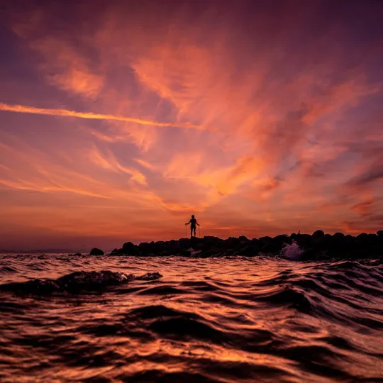 Person stood on rocks by sea and colourful sky above