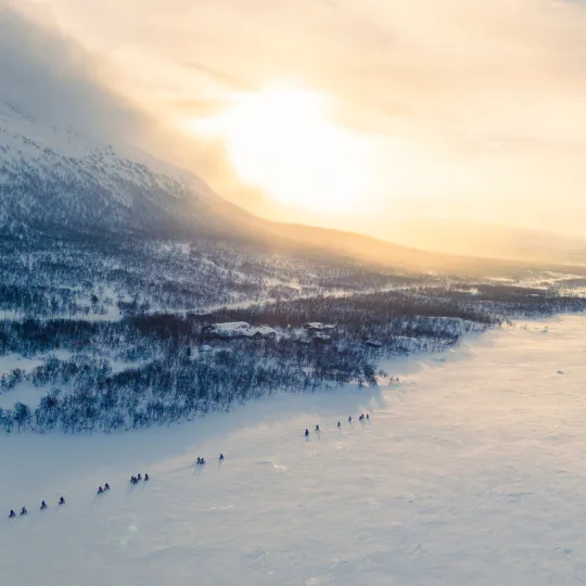 Aerial view of cyclists riding across a frozen snow-covered landscape at sunrise, surrounded by mountains and soft winter light.