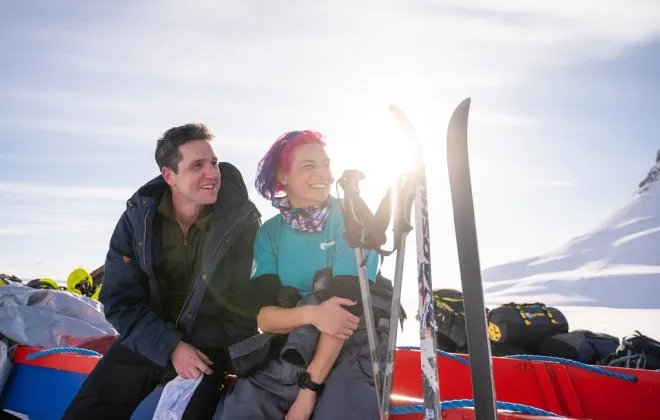 Woman with colourful hair and man sitting beside skis in a snowy mountain setting and both smiling