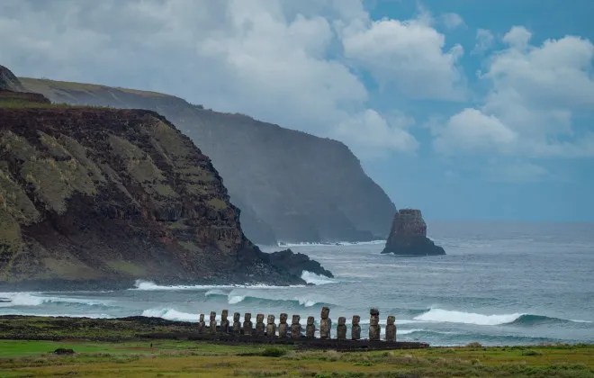 Statues stood infront of crashing waves and a mountainous terrain, with blue sky and clouds behind
