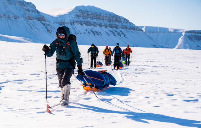 Participants pulling sleds through snow with a bright sky above them and snow-covered mountains in the background